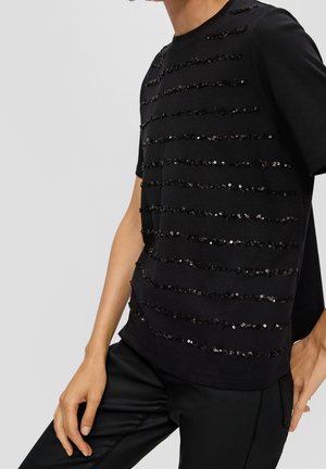 Personne portant un haut noir à manches courtes avec des rangées horizontales de sequins noirs et un pantalon noir, debout devant un fond clair uni.