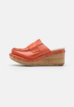 Zueco de cuero naranja sin cordones con tacón de cuña de madera y tachuelas plateadas alrededor de la suela, exhibido sobre un fondo blanco.