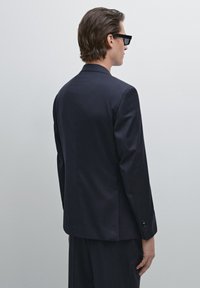 Marineblaues Jacket mit taillierter Passform, mit einem Schlitzt hinten und strukturiertem Stoff. Es hat lange Ärmel und Knopfmanschetten, kombiniert mit passenden Hosen.