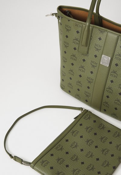 Green leather tote bag and matching pouch with black MCM logo pattern and silver hardware on a white surface.