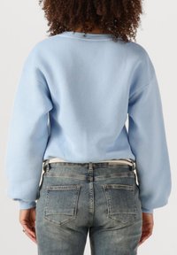 Person mit lockigem Haar, der einen hellblauen Sweatshirt und eine ausgewaschene blå Jeans trägt, von hinten vor einem schlichten Hintergrund gezeigt.