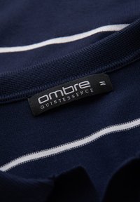 Námořnická modrá košile s bílými vodorovnými pruhy, s černým štítkem označeným "ombre QUINTESSENCE" a velikostí "M" blízko límce.