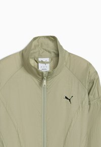 Veste légère vert olive clair avec fermeture éclair, col montant et logo Puma noir sur la poitrine gauche. Tissu texturé avec des coutures visibles.