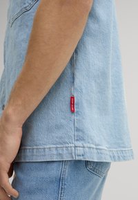 Camicia di jeans azzurro chiaro con maniche corte, con etichetta rossa lungo la cucitura laterale e dettagli di cucitura in vista.