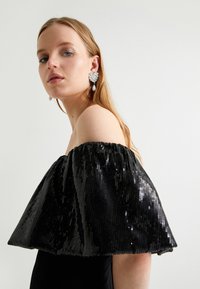 Top noir à paillettes, sans manches, avec un design de manches volumineuses et un corps lisse. Boucles d'oreilles argentées originales avec des perles.