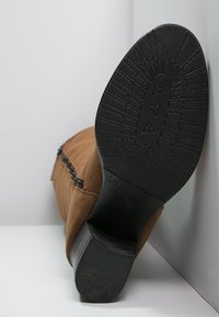 Botín de ante marrón con suela de goma, que presenta un tacón negro texturizado y una cremallera lateral. La parte inferior muestra un diseño de banda de rodadura con patrón.