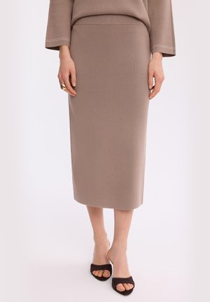 Kobieta nosząca beżową midi ołówkową spódnicę, dopasowany top, czarne buty na otwartym obcasie oraz złoty efektowny pierścionek na lewej ręce.