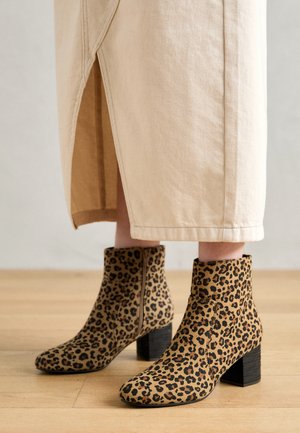 Bottines à imprimé léopard avec talons blocs portées avec un pantalon beige à jambes larges avec fentes latérales, debout sur un sol en bois.
