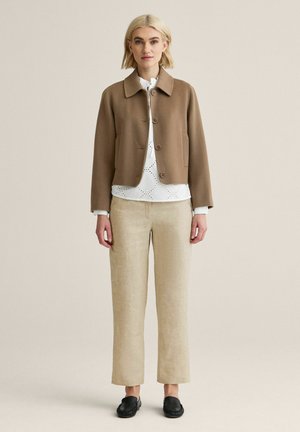 Lyshåret kvinde iført en brun knappet jakke over en hvid bluse med mønster, beige bukser med lige ben og sorte slip-on sko.