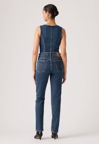 Γυναίκα με τα μαλλιά της σε κότσο, που φοράει αμάνικο τοπ από τζιν και ψηλόμεσο, ίσιο τζιν, στέκεται με την πλάτη προς την κάμερα.