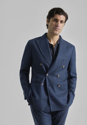Chaqueta de traje azul marino de doble botonadura, confeccionada en tela texturizada con grandes botones, combinada con una camisa azul marino de diseño ajustado.