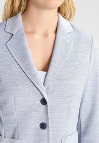 Blazer rayé bleu clair avec des revers, deux boutons sur le devant et des poches latérales, fabriqué dans un tissu texturé.