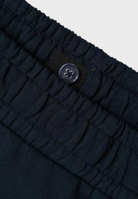 Ceinture élastique marine avec texture froncée, dotée d'un bouton rond en plastique transparent. Vue en gros plan mettant en valeur le détail du tissu et des fermetures.