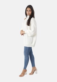 Femme debout portant un pull en tricot blanc à épaules dénudées, un jean skinny bleu et des sandales à talons transparents, les mains jointes devant.