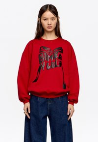 Młoda kobieta w czerwonym sweatshirtie z czarną grafiką kokardy i napisem, w luźnych ciemnoniebieskich dżinsach, stojąca na tle białym.