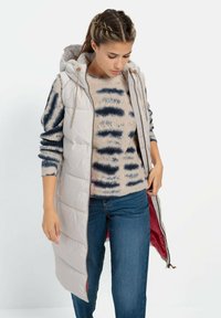 Lys grå polstret vest med hætte, som har en frontlynlås og snore. Båret over en beige mønstret sweater og jeans.