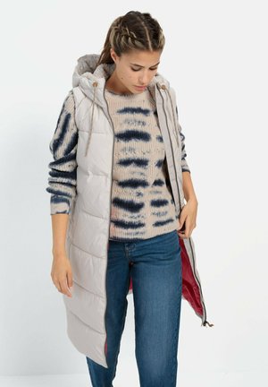 HellgraWest-Weste mit Kapuze, ausgestattet mit einem Reißverschluss auf der Vorderseite und Kordelzügen. Getragen über einem beigen gemusterten Pullover und Jeans.