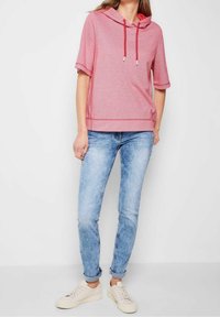 Kvinde iført en pink kortærmet hættetrøje med røde snore, lyseblå opsmøgede jeans og hvide sneakers, stående foran en hvid baggrund.