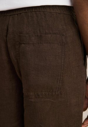 Közeli kép sötétbarna, texturált szövetnadrágról, amelynek négyzet alakú hátsó zsebe és látható varrásai vannak egy személy csípőjén és kezén.