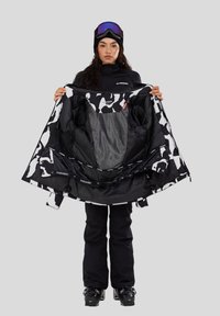 Frau in schwarzer Skibekleidung und Skibrille hält eine schwarz-weiß gemusterte Skijacke geöffnet und zeigt das Innenfutter.