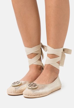 Zapatos planos metálicos dorados con suelas de yute trenzado, que presentan un acento de logo redondo en la puntera y correas beige en cruz alrededor de los tobillos.
