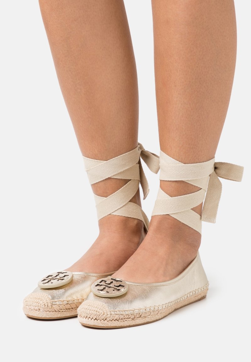 Zapatos planos metálicos dorados con suelas de yute trenzado, que presentan un acento de logo redondo en la puntera y correas beige en cruz alrededor de los tobillos.