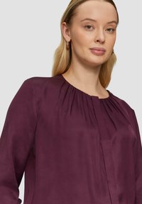 Burgunderfarbene Bluse mit langen Ärmeln, weicher Textur, gerafftem Detail am Kragen und einer Frontleiste zum Verschließen. Minimalistisches Design.