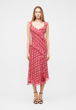 Žena v ružových a červených vzorovaných midi šatách bez rukávov s volánikmi, stojaca vzpriamene pred jednoliatym bielym pozadím.