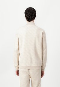 Sweatshirt beige à motifs avec un col haut, des poignets et un ourlet côtelés. Texture douce avec un design géométrique sur l'ensemble.