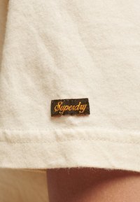 Tela de algodón beige con un acabado texturizado y una etiqueta negra que presenta la palabra "Superdry" en un bordado dorado en cursiva cerca del dobladillo.