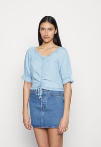 Blusa curta azul clara com mangas bufantes, decote com pregas e cintura com cordão, combinada com uma saia mini de ganga. Textura suave.