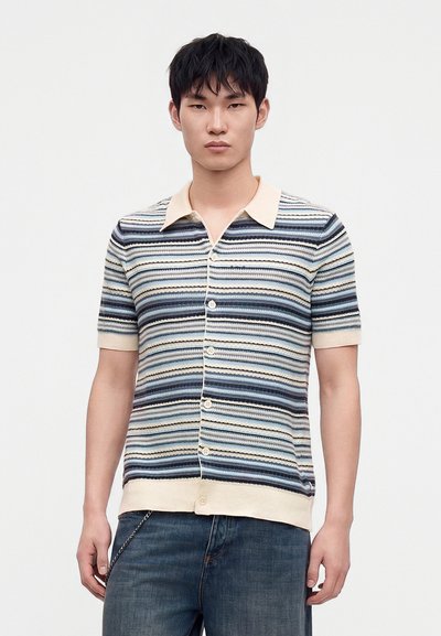 Jeune homme portant une chemise en maille à manches courtes boutonnée avec des rayures horizontales bleues, blanches et beiges et un jean bleu sur fond blanc.