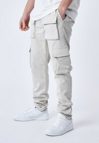 Pantaloni cargo beige con più tasche laterali e una vestibilità affusolata, abbinati a sneakers bianche da ginnastica. Tessuto dalla superficie liscia.