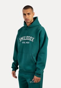 Felpa in cotone verde con tasca a marsupio frontale, polsini e orlo a coste. Presenta la scritta "SMILODOX ATHL WEAR" in un testo bianco a contrasto.