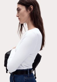 Weißes, geripptes Langarmoberteil mit Rundhalsausschnitt, figurbetonter Form, kombiniert mit hochgeschnittenen Jeans und einem schwarzen Rucksack.