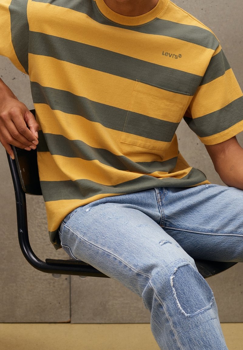 Personne portant un t-shirt rayé jaune et gris Levi's et un jean bleu clair déchiré, assise sur une chaise noire contre un mur gris.