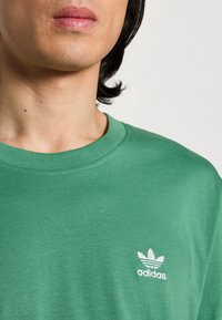 Grön bomullströja med rund halsringning och en vit broderad Adidas-logga på vänster bröst. Slät textur, avslappnad passform.