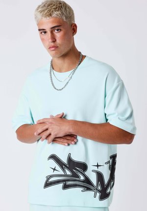 Giovane uomo con capelli corti decolorati indossa una T-shirt oversize azzurro chiaro con stampa graffiti nera sull'orlo e collane a catena argentate.