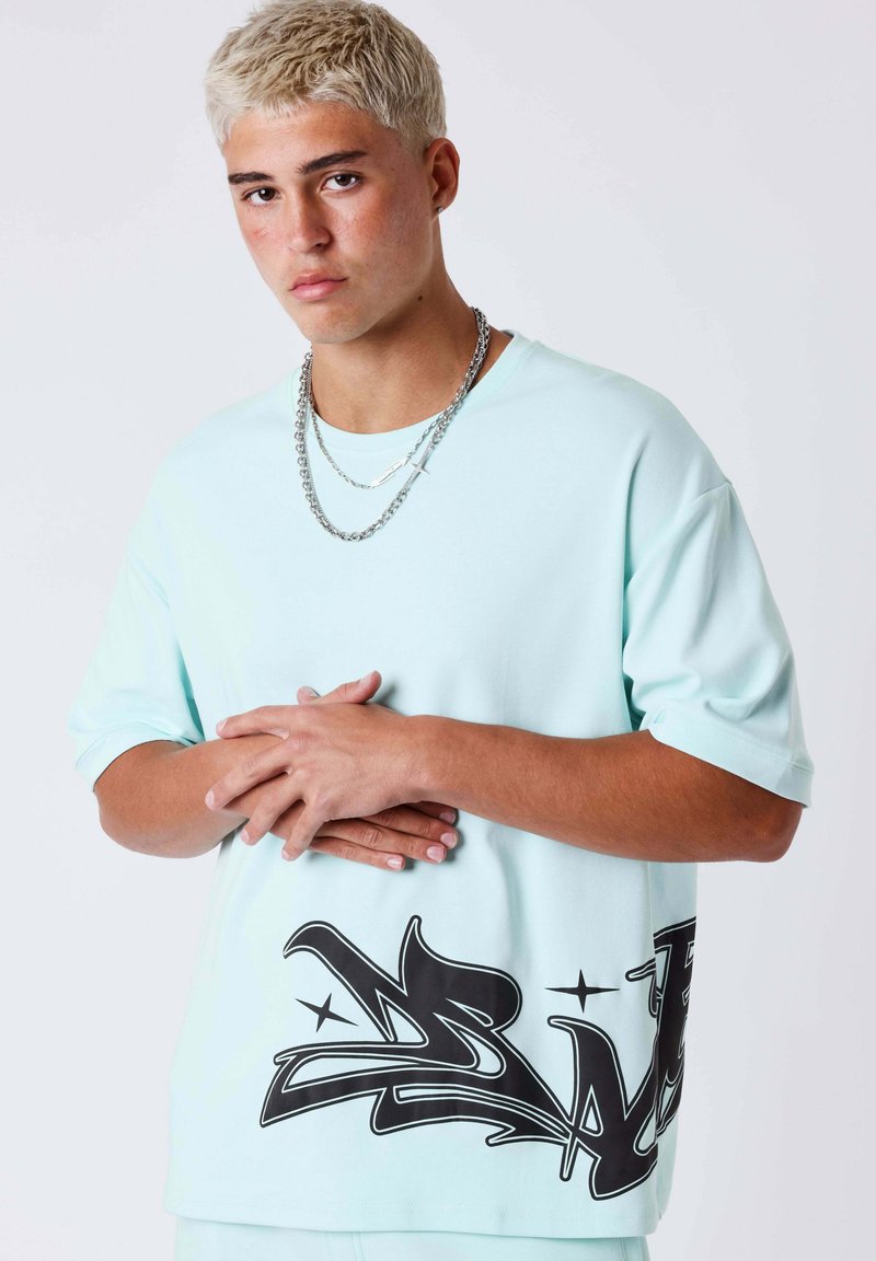 Jeune homme aux cheveux courts et décolorés portant un T-shirt bleu clair oversize avec un imprimé graffiti noir à l'ourlet et des chaînes en argent autour du cou.
