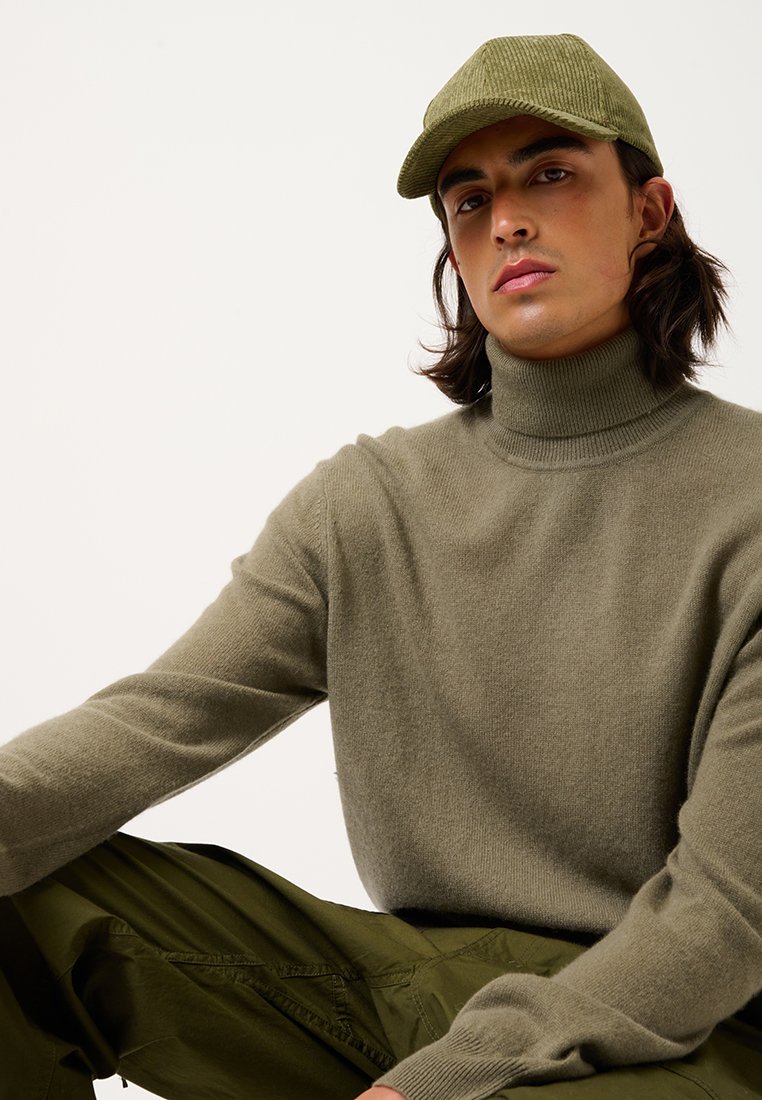 Mann mit langen Haaren, der eine olivgrüne Cordmütze, einen passenden Rollkragenpullover und olivgrüne Hosen trägt, sitzt vor einem schlichten Hintergrund.