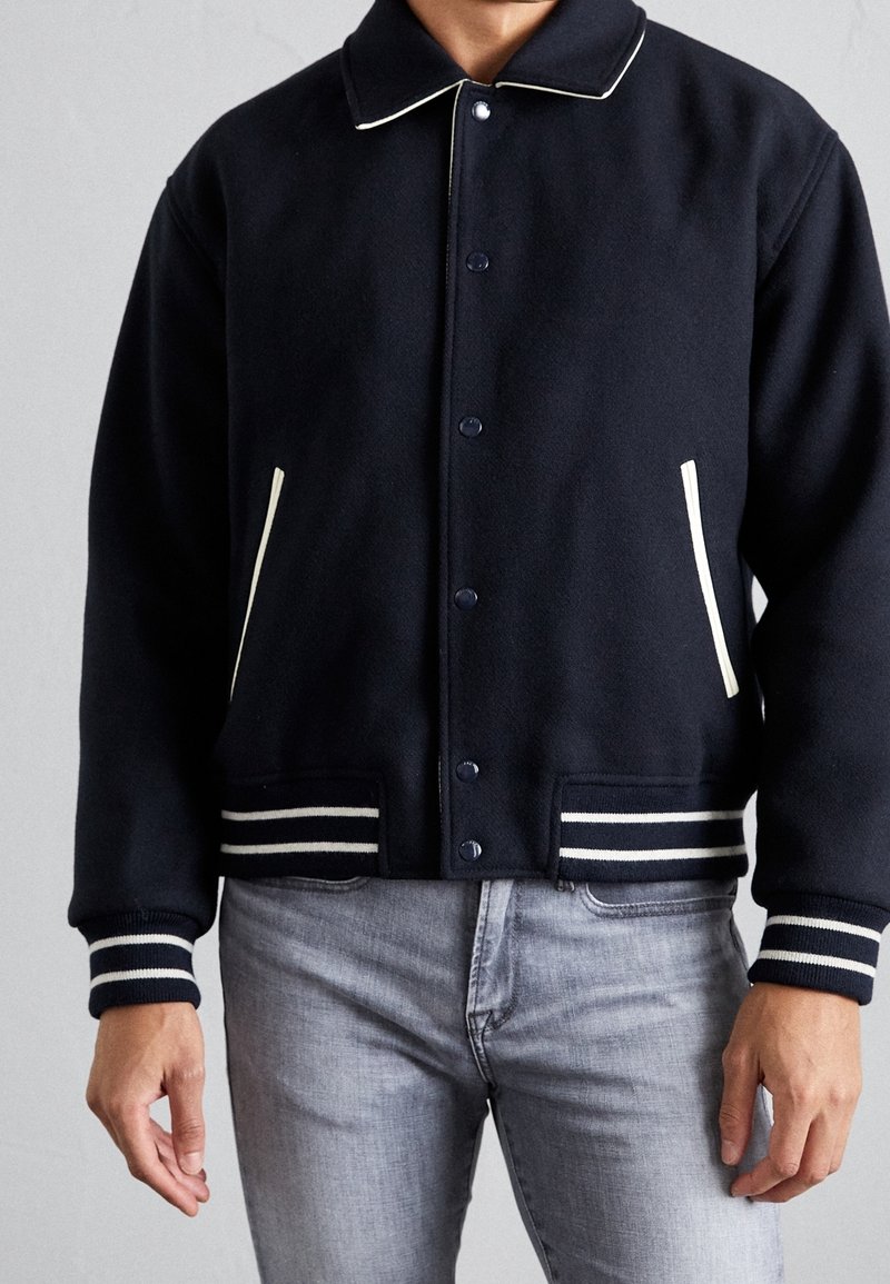 Homme portant une veste style varsity noire avec des poignets et un ourlet rayés blancs, des poches bordées de blanc et un jean gris clair.