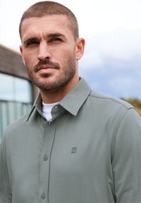 Ljusgrön skjorta med krage och slät textur, knappstängning fram och subtil logotyp. Bärs över en vit skjorta. Utomhusbakgrund.