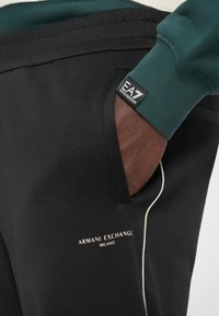 Tuvumā redzama roka melnās Armani Exchange biksēs ar baltu apdari un EA7 Emporio Armani logotipu uz zaļas piedurknes.