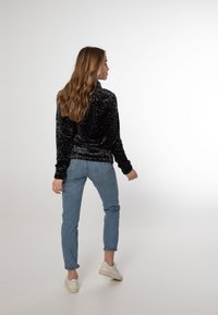 Giubbotto di felpa nero con stampa leopardata, colletto alto, abbinato a jeans azzurri e sneaker bianche, visto da dietro.