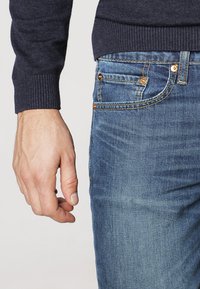 Dark navy sweater arm next to light blue denim jeans. Jeans feature copper hardware, contrast stitching, and a slim fit design.