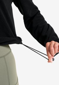 Svart fleecepullover med justerbara dragsnören i nederkant, med slät textur och avslappnad passform, matchad med olivgröna byxor.