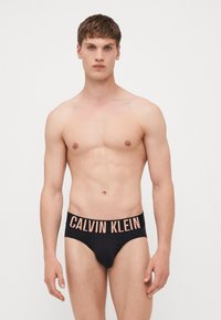 Boxers en coton noir avec une large ceinture élastique mettant en avant "CALVIN KLEIN" en lettres contrastantes orange clair. Texture lisse, design classique.