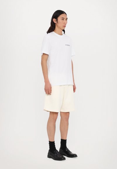 T-shirt en coton blanc à manches courtes avec un petit graphique sur la poitrine, associée à un short crème et des chaussures noires à enfiler.