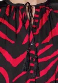 Blouse noire à larges rayures zébrées rouges, col lacé en cordon noir avec pompons, et boutons noirs sur le devant.