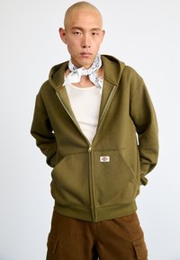 Sweatshirt verde-azeitona com fecho éclair, feito de fleece suave, com bolsos tipo canguru, punhos canelados e um patch com a marca no bolso esquerdo.